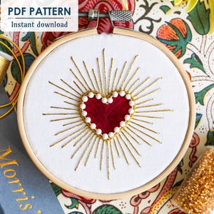 May include: A white embroidery hoop featuring a red heart surrounded by gold thread and white beads. The heart is stitched with a radiating pattern of gold thread. The hoop is on a floral patterned fabric.