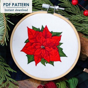 May include: A red poinsettia flower embroidery pattern in a wooden embroidery hoop. The hoop is surrounded by green pine branches and red berries.