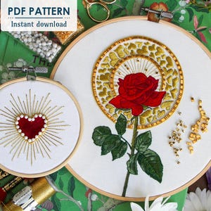 Könnte beinhalten: Zwei Stickmuster in Stickrahmen, eines mit einem roten Herzen, das von goldenen Strahlen umgeben ist, und das andere mit einer roten Rose mit goldenen Blättern und einem goldenen Herzmuster dahinter.