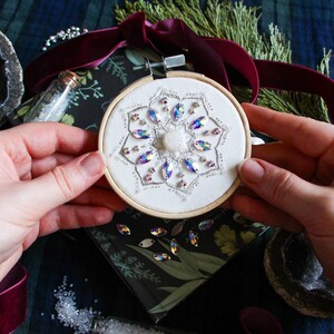 My own hands holding the finished embroidery to indicate scale. The hoop is quite small and is suitable for use as a Christmas ornament. The hoop is being held over a black book and is surrounded by decorative objects in the background.