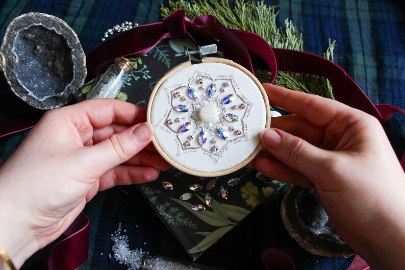 My own hands holding the finished embroidery to indicate scale. The hoop is quite small and is suitable for use as a Christmas ornament. The hoop is being held over a black book and is surrounded by decorative objects in the background.