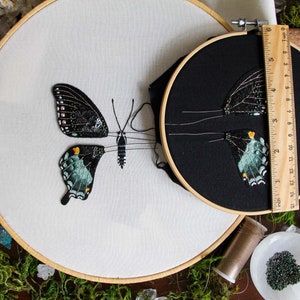 2 embroidery hoops of the 3D butterfly before assembly. One 6 inch hoop with the butterfly wings on black fabric, and a larger hoop with the butterfly body on white fabric and cut out left wings. Styled with moss and embroidery materials.