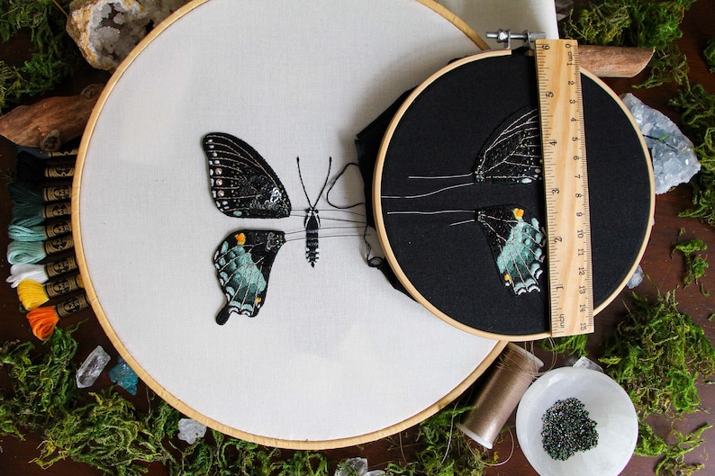 2 embroidery hoops of the 3D butterfly before assembly. One 6 inch hoop with the butterfly wings on black fabric, and a larger hoop with the butterfly body on white fabric and cut out left wings. Styled with moss and embroidery materials.