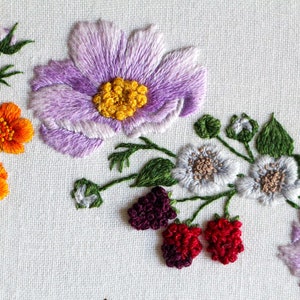 A close up of the embroidered pale purple cosmos, grey blackberry flowers with purple blackberries, and bright orange buttercup flowers embroidered using thread painting techniques on a white fabric ground.