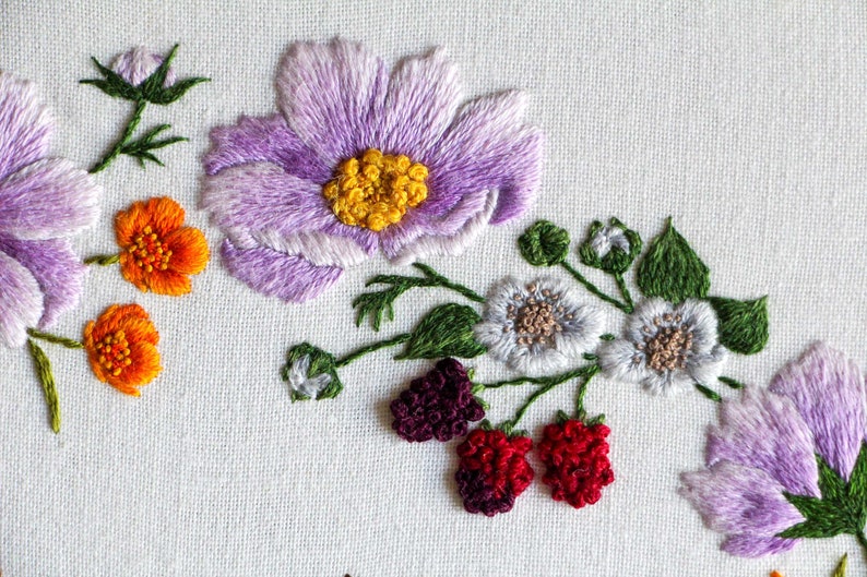 A close up of the embroidered pale purple cosmos, grey blackberry flowers with purple blackberries, and bright orange buttercup flowers embroidered using thread painting techniques on a white fabric ground.