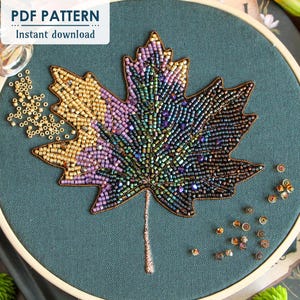 Puede incluir: Primer plano de un aro de bordado con cuentas con un diseño de hoja de arce de colores. La hoja está hecha de varios tonos de cuentas moradas, verdes, doradas y marrones. El aro está sobre un fondo de tela verde oscuro.