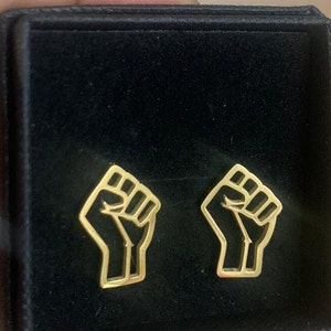 May include: Gold-coloured fist-shaped earrings presented in a black jewellery box. The earrings feature a cutout design, highlighting the fist's outline. The jewellery box is closed, with the earrings inside.