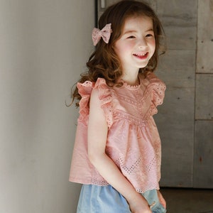 Op de afbeelding: Een jong meisje draagt een roze geborduurde top met een kantversiering en een blauwe denim rok met een geruite zoom. Ze glimlacht en heeft een roze strik in haar haar.