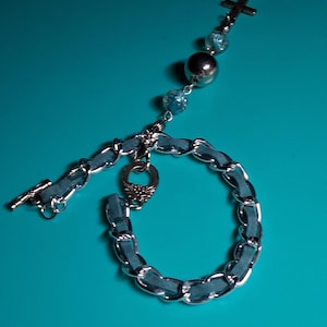 May include: A silver-toned bracelet with a blue chain-like design. The bracelet features a cross charm, blue beads, and a toggle clasp. The background is a solid teal color, highlighting the jewelry.