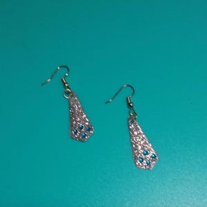May include: A pair of silver-toned dangle earrings. Each earring has a triangular shape with a textured surface and small blue gemstones. The earrings hang from a silver-colored hook.