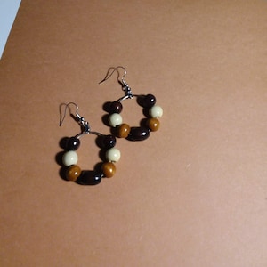 May include: A pair of hoop earrings with a mix of brown, tan, and cream-colored beads. The earrings have silver-colored ear wires and are displayed on a brown surface.