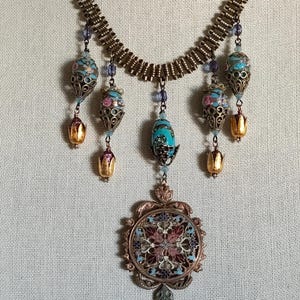May include: A gold and bronze necklace with a large, round pendant featuring a colorful mosaic design. The pendant is surrounded by smaller, dangling charms with turquoise, pink, and gold accents.