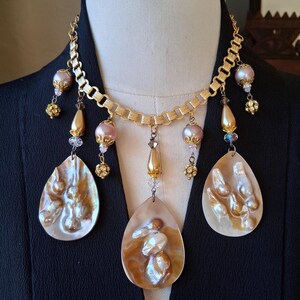 May include: A gold-toned necklace with a chain design, featuring teardrop-shaped pendants with a pearlescent finish. The necklace includes pink and gold beads, and crystal accents. The necklace is displayed on a mannequin.