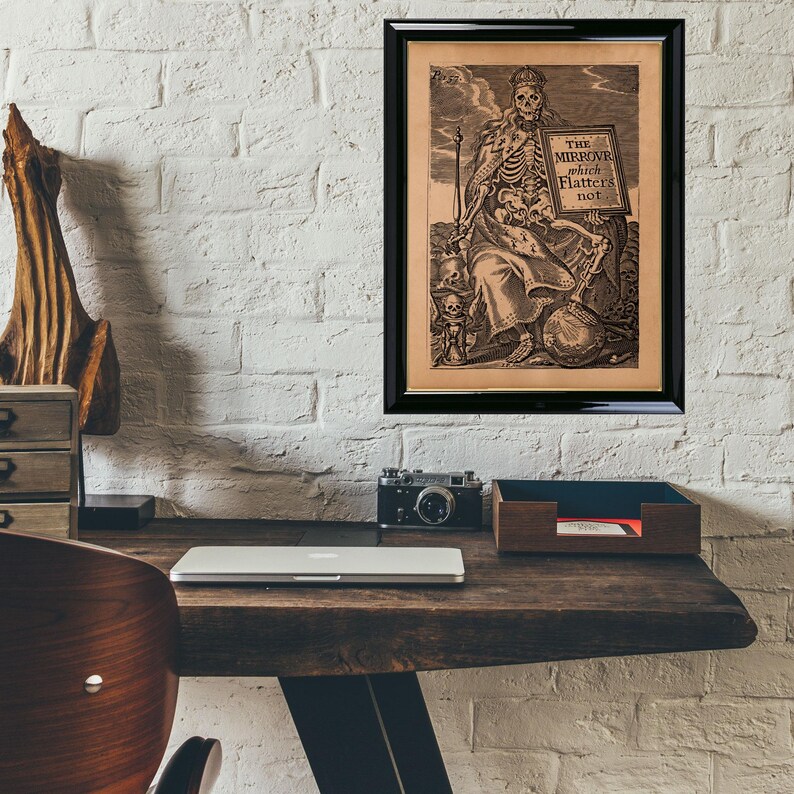 May include: A framed sepia-toned print in a black frame, featuring a skeleton holding a sign with the text "THE MIRROVR which flatters not." The artwork is displayed on a white brick wall.