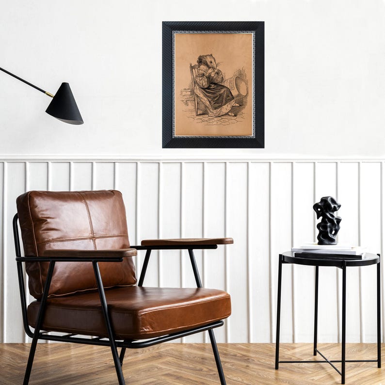 K&ouml;nnte beinhalten: Gerahmte Vintage-Illustration einer sitzenden Figur mit einem nagetierartigen Gesicht, die ein Kleid tr&auml;gt und ein Buch h&auml;lt. Der schwarze Rahmen hat einen strukturierten Rand. Das Bild h&auml;ngt an einer wei&szlig;en Wand &uuml;ber einem braunen Ledersessel und einem schwarzen Beistelltisch.