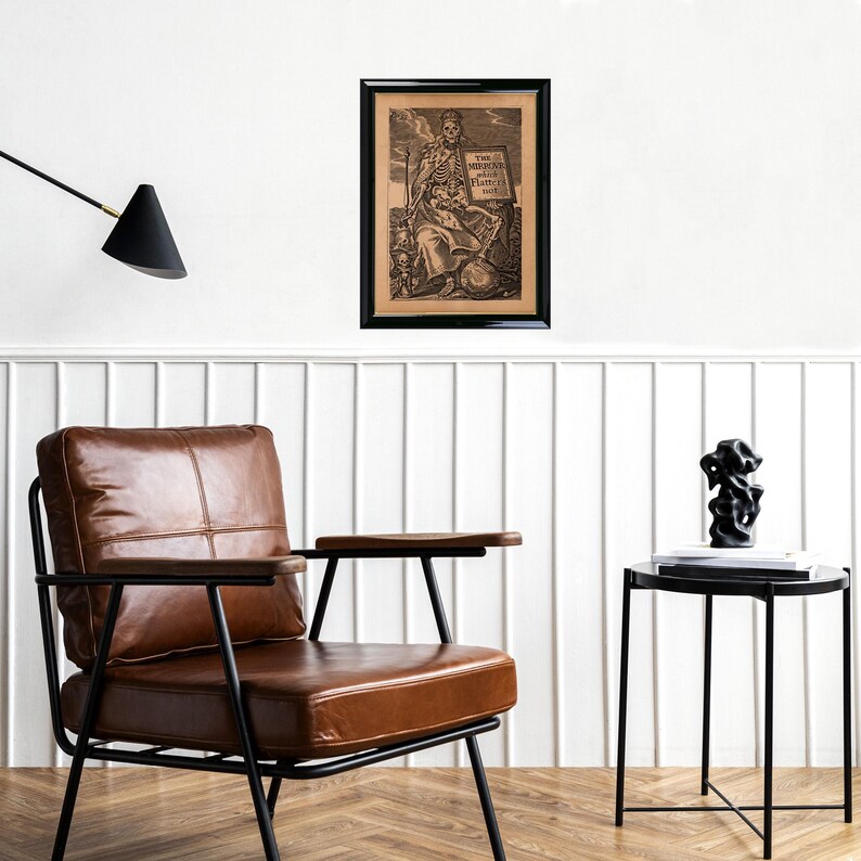 May include: A framed monochrome print featuring a skeleton figure holding a sign. The artwork hangs on a white wall above a brown leather armchair and a black side table with a sculpture.