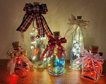 May include: Several glass bottles with cork stoppers, each filled with fairy lights. The bottles are decorated with colourful ribbons and bows, creating a festive display. The bottles are arranged on a wooden surface, with a warm, inviting glow.