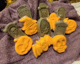 May include: A collection of Halloween-themed bath bombs in various shapes, including tombstones, pumpkins, ghosts, bats, and skulls. The bath bombs are orange and gray, wrapped in clear plastic, and arranged on a purple towel.