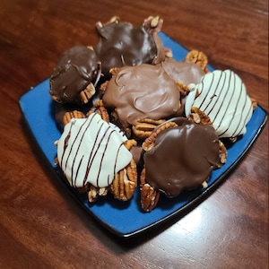 May include: A blue square plate holds a collection of chocolate-covered pecan candies. Some are coated in dark chocolate, others in milk chocolate, and a few are drizzled with white chocolate. The candies sit atop a dark wooden surface.