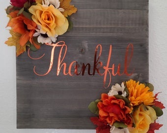 May include: A square wooden sign with a gray finish, adorned with the word "Thankful" in copper-colored script. The sign is decorated with colorful fall-themed floral arrangements in the corners, including orange, yellow, and red flowers.