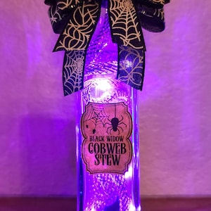 May include: A rectangular glass bottle with a cork stopper, filled with purple lights. A black and white spiderweb bow adorns the top. The label reads "Black Widow Cobweb Stew."