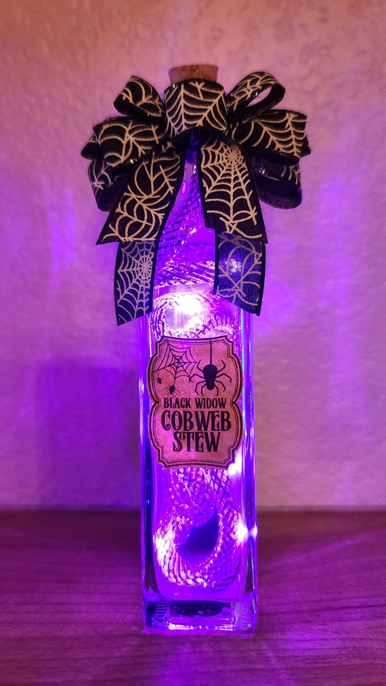 May include: A rectangular glass bottle with a cork stopper, filled with purple lights. A black and white spiderweb bow adorns the top. The label reads "Black Widow Cobweb Stew."