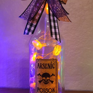 May include: A clear glass bottle with a cork stopper, decorated for Halloween. It features a label that reads "Arsenic Poison - Do Not Drink" and is adorned with orange lights and a decorative bow. The bow has purple and black accents.