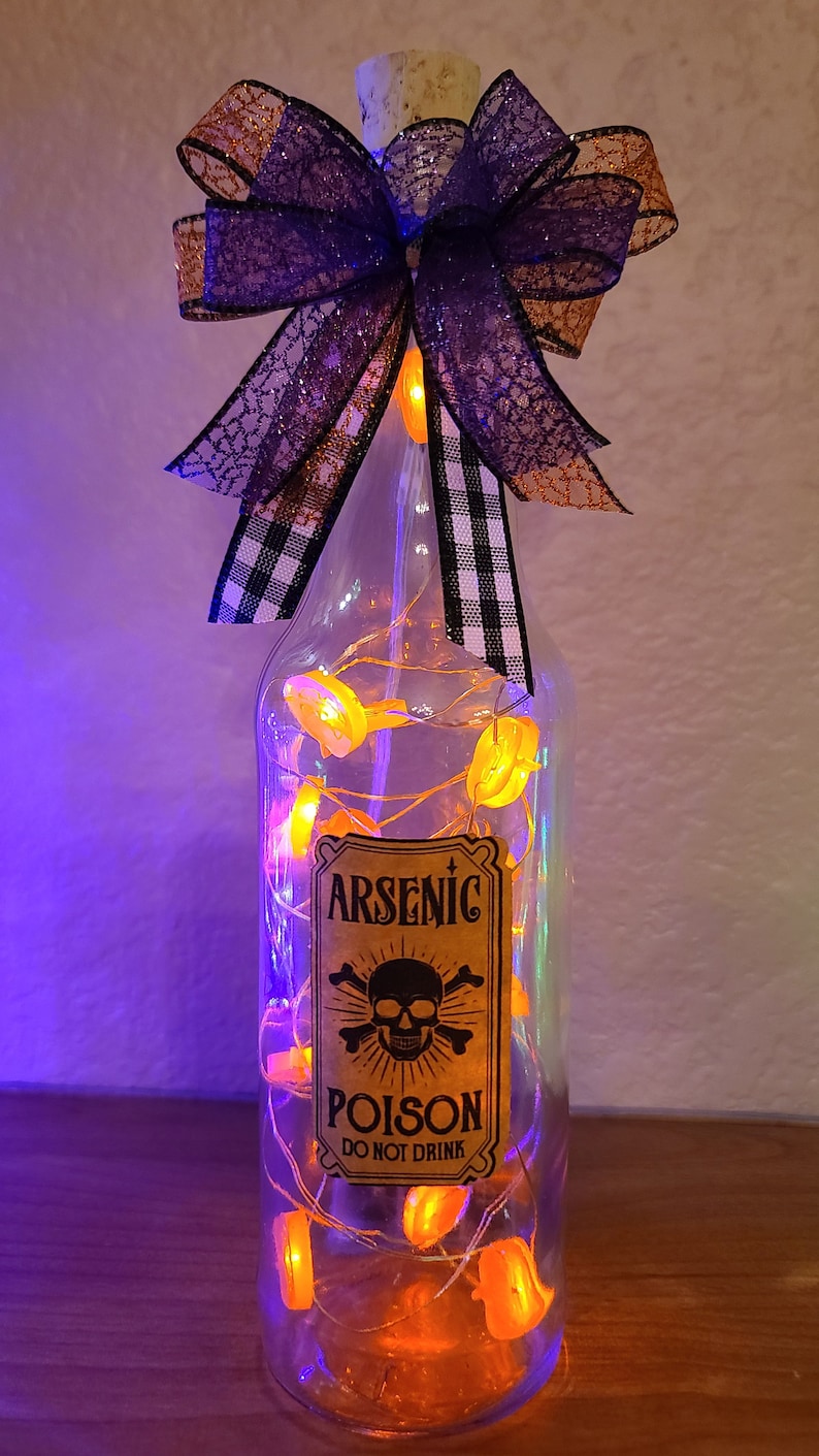 May include: A clear glass bottle with a cork stopper, decorated for Halloween. It features a label that reads "Arsenic Poison - Do Not Drink" and is adorned with orange lights and a decorative bow. The bow has purple and black accents.