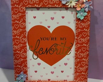 May include: A rectangular red picture frame with a repeating "love" and heart pattern. The frame holds a card with a large red heart and the gold text "YOU'RE MY favorite". The frame is decorated with small, colorful paper flowers.