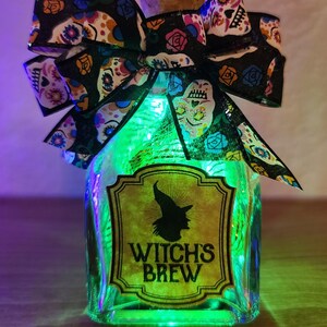 May include: A decorative glass bottle labeled "Witch's Brew" with a witch silhouette. The bottle is adorned with a black bow featuring colorful sugar skull designs. The bottle is lit with green fairy lights, creating a festive Halloween theme.
