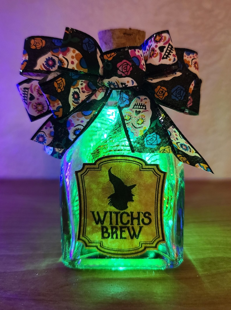 May include: A decorative glass bottle labeled "Witch's Brew" with a witch silhouette. The bottle is adorned with a black bow featuring colorful sugar skull designs. The bottle is lit with green fairy lights, creating a festive Halloween theme.