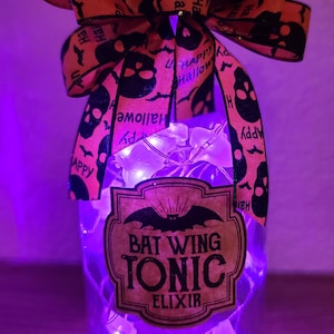 May include: A clear glass bottle with a cork stopper, adorned with a Halloween-themed orange and black bow. The bottle features a label that reads "Bat Wing Tonic Elixir" and is illuminated by fairy lights.
