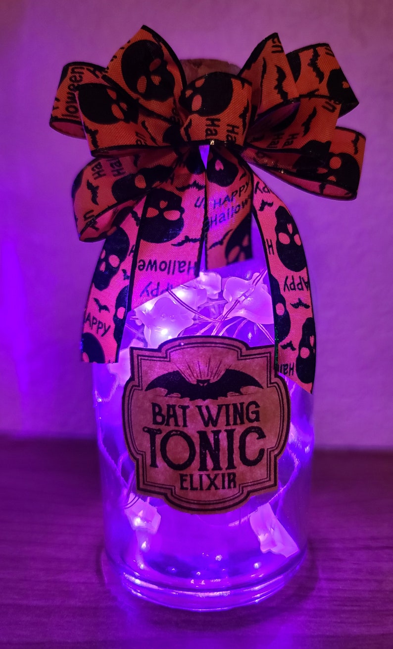 May include: A clear glass bottle with a cork stopper, adorned with a Halloween-themed orange and black bow. The bottle features a label that reads "Bat Wing Tonic Elixir" and is illuminated by fairy lights.