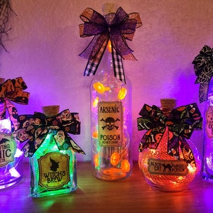 Puede incluir: Botellas decorativas con temática de Halloween con tapones de corcho y lazos festivos. Cada botella presenta una etiqueta única con textos como "Arsenic Poison" y "Bat Wing Tonic Elixir". Las botellas están iluminadas con luces de hadas, creando un ambiente espeluznante.