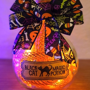 May include: A clear glass bottle with a cork stopper, filled with orange lights and a decorative bow. The bow is black with Halloween-themed patterns. A label reads "Black Cat Magic Potion" with a black cat silhouette.
