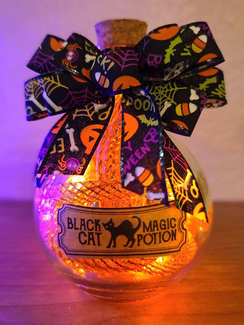 May include: A clear glass bottle with a cork stopper, filled with orange lights and a decorative bow. The bow is black with Halloween-themed patterns. A label reads "Black Cat Magic Potion" with a black cat silhouette.