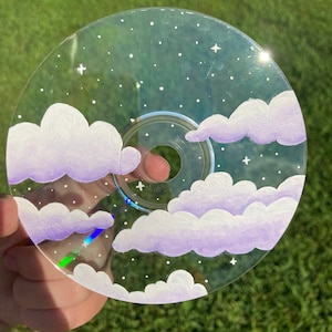 Peut inclure: Un CD transparent avec un motif peint de nuages blancs avec des accents violets et des étoiles blanches sur un fond bleu.