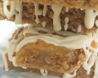 May include: A stack of three homemade granola bars with a white chocolate drizzle. The bottom bar has a layer of caramel visible.