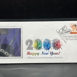 May include: A commemorative cachet envelope with a silver 2000 design and a stamp featuring a Times Square 2000 Station image. The envelope also features a silver 2000 design with fireworks and the text "Happy New Year!" and "Official Commemorative Cachet".
