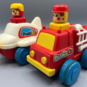 May include: Two colorful toy vehicles: a red and yellow fire truck and a white and red airplane. Both toys have blue wheels and a cartoon figure on top. The fire truck has a white ladder and the airplane has a yellow propeller. The text "Push'n Go" is on both toys.