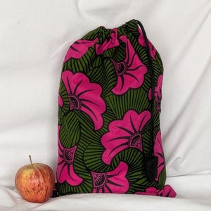 Könnte beinhalten: Ein Kordelzugbeutel mit einem lebendigen Muster aus großen rosa Blumen auf dunkelolivgrünem Hintergrund mit schwarzen Liniendetails. Der Beutel ist oben mit einer schwarzen Kordel und einem schwarzen Stopper gerafft und steht neben einem rot-gelben Apfel.