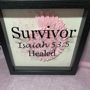 May include: A framed artwork featuring a pink ribbon flower design. The word "Survivor" is displayed in black script, along with the text "Isaiah 53:5 Healed". Pink ribbon accents are scattered across the background.
