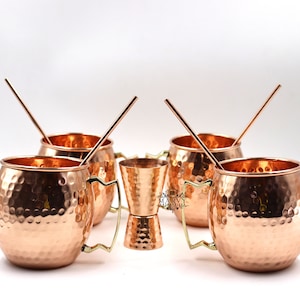 Könnte beinhalten: Set aus Kupfer-Moscow-Mule-Bechern mit gehämmertem Muster, goldfarbenen Griffen und Kupferstrohhalmen. Ein Kupfer-Jigger ist ebenfalls enthalten. Die Becher stehen auf einer weißen Oberfläche, ideal für Cocktails.