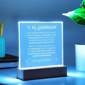 May include: A light blue illuminated acrylic plaque with a wooden base. The plaque has a message for a grandson, "To My GRANDSON As you journey through life, you'll encounter numerous challenges. Embrace them and give them your all. Remember, life isn't about waiting for the storm to pass; it's about standing strong and thriving in the rain. While not every day may be perfect, seek out something good in each one. Laugh, love, and truly live. Chase after your dreams, have unwavering belief in yourself, and never forget to RADIATE AWESOMENESS. Love you forever"