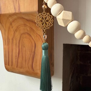 May include: A decorative wooden bead garland with a gold filigree charm, a clear crystal, and a green tassel. The garland features various shapes of wooden beads, including spheres and a geometric shape.