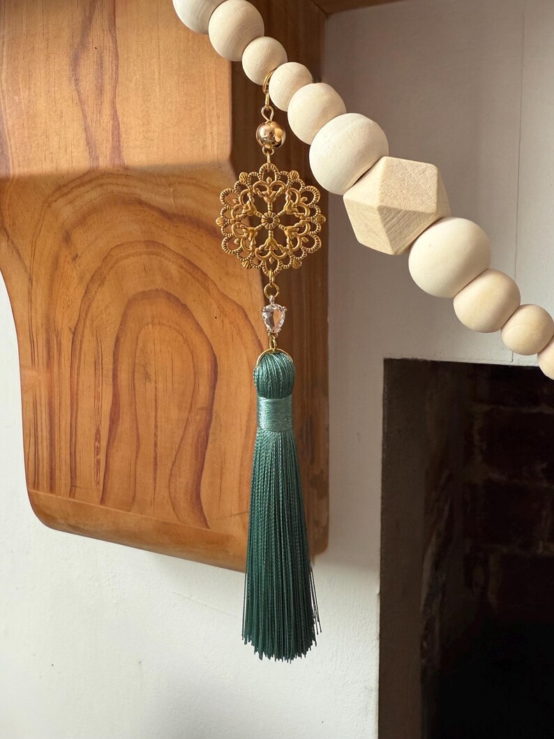 May include: A decorative wooden bead garland with a gold filigree charm, a clear crystal, and a green tassel. The garland features various shapes of wooden beads, including spheres and a geometric shape.