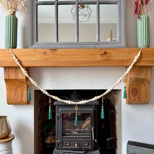 May include: A decorative garland of off-white beads with gold accents and teal tassels hangs above a fireplace. The fireplace has a black stove, a wooden mantel, and a gray framed mirror. Two green vases with dried flowers sit on the mantel.