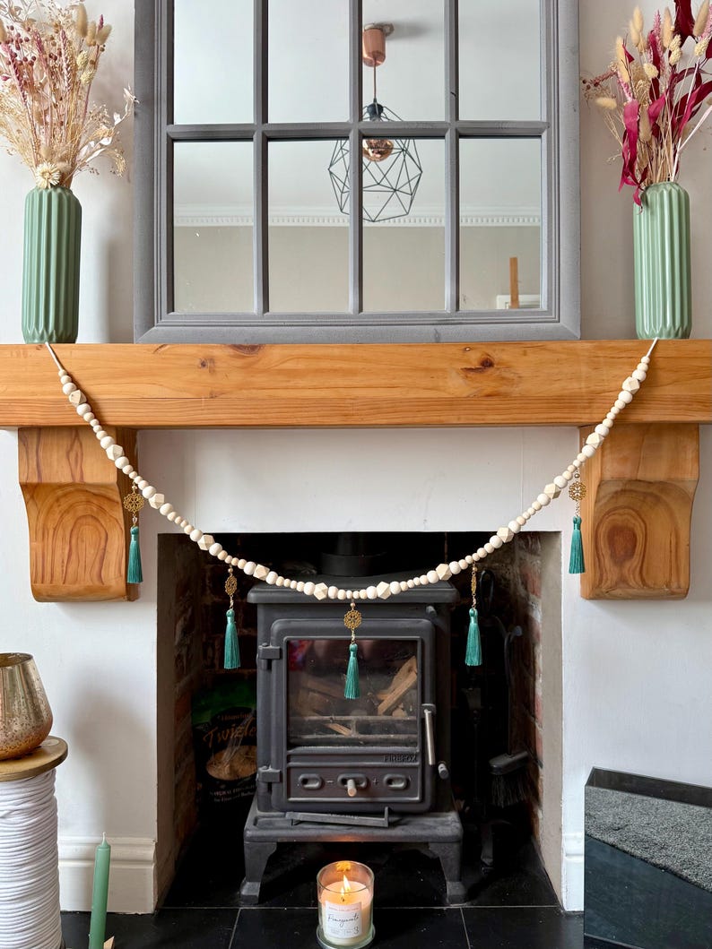 May include: A decorative garland of off-white beads with gold accents and teal tassels hangs above a fireplace. The fireplace has a black stove, a wooden mantel, and a gray framed mirror. Two green vases with dried flowers sit on the mantel.
