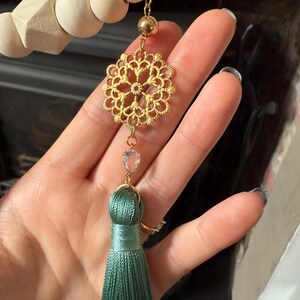 May include: A gold filigree pendant with a clear crystal heart and a teal tassel. The pendant is suspended from a wooden bead garland. The tassel is made of silky threads.