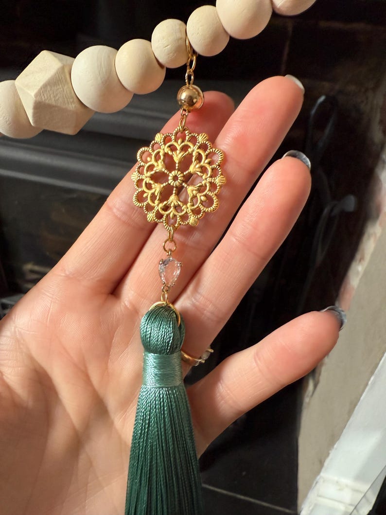 May include: A gold filigree pendant with a clear crystal heart and a teal tassel. The pendant is suspended from a wooden bead garland. The tassel is made of silky threads.